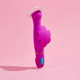The Aria Rechargeable Vibe Naughty AF-Plum by Blush Novelties stands upright with a butterfly accent and teal button, topped with a small white cowboy hat. Made from Puria platinum-cured silicone, it's shown against a pink background.